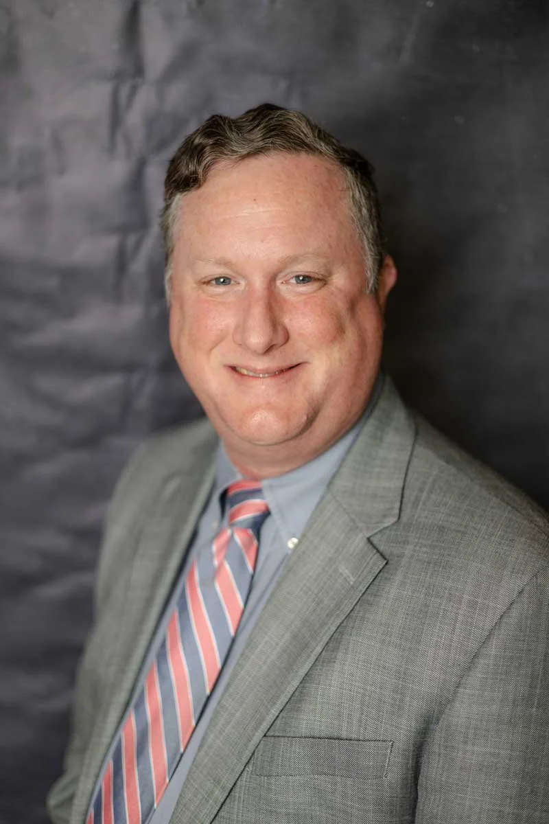 A professor at The Cambridge School of Dallas classical Christian school grades 5-12, with short, wavy hair wearing a gray suit, light blue shirt, and pink striped tie.