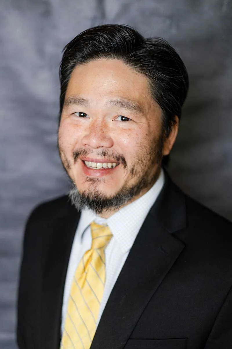 A professor at The Cambridge School of Dallas classical Christian school grades 5-12, wearing a black suit, white shirt, and yellow striped tie, against a dark gray background.