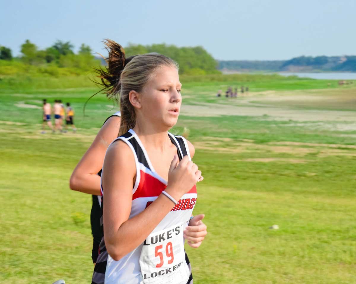 A young girl running outdoors on a grassy field during daytime, participating in a race, with a river and trees in the background.