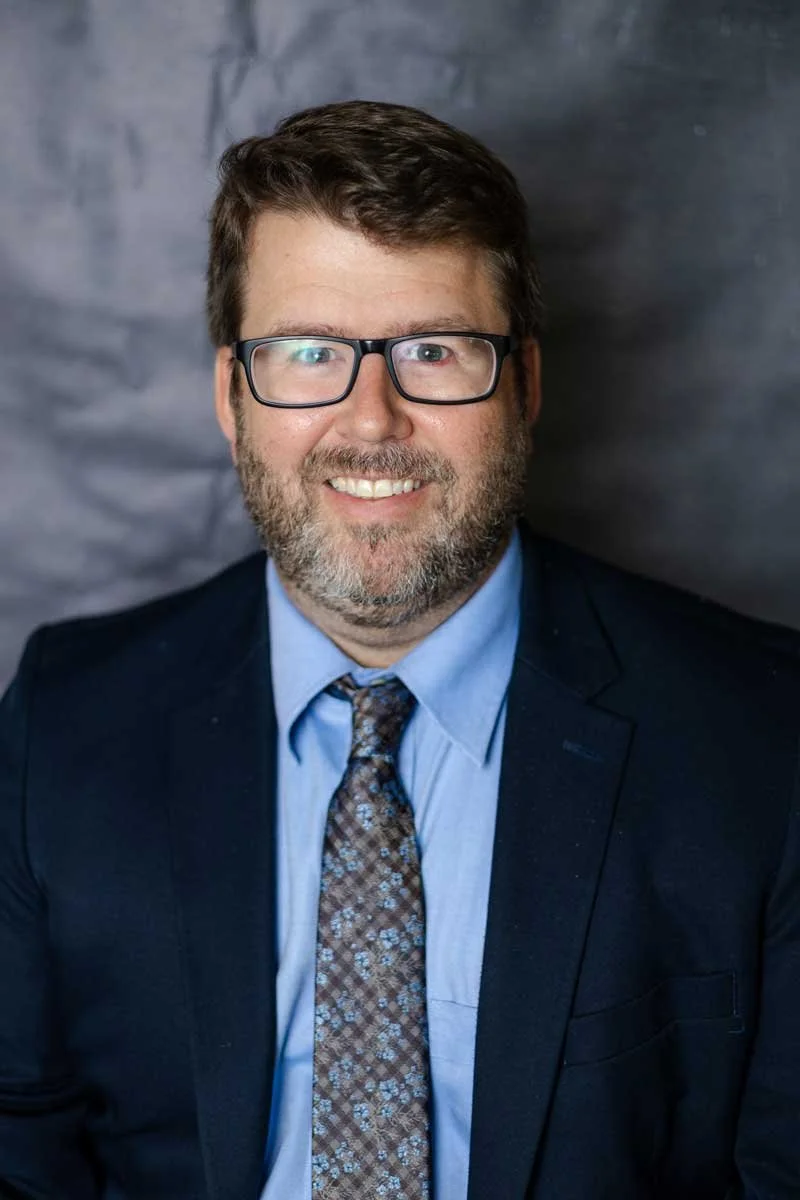A professor at The Cambridge School of Dallas classical Christian school grades 5-12. A portrait of a man with glasses, brown hair, and a beard, wearing a dark suit, light blue shirt, and patterned tie, smiling in front of a gray backdrop.