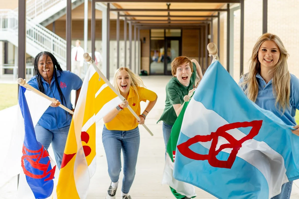 Four students who are House Leaders at the Cambridge School of Dallas running outside a school holding colorful House flags and smiling.