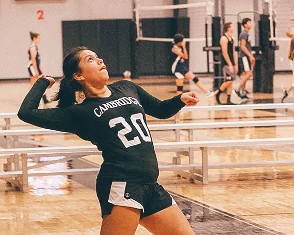 A young female volleyball player in a black and white uniform with number 20 and the word 'Cambridge' on her jersey is preparing to serve the volleyball in an indoor gymnasium, with other players practicing in the background.