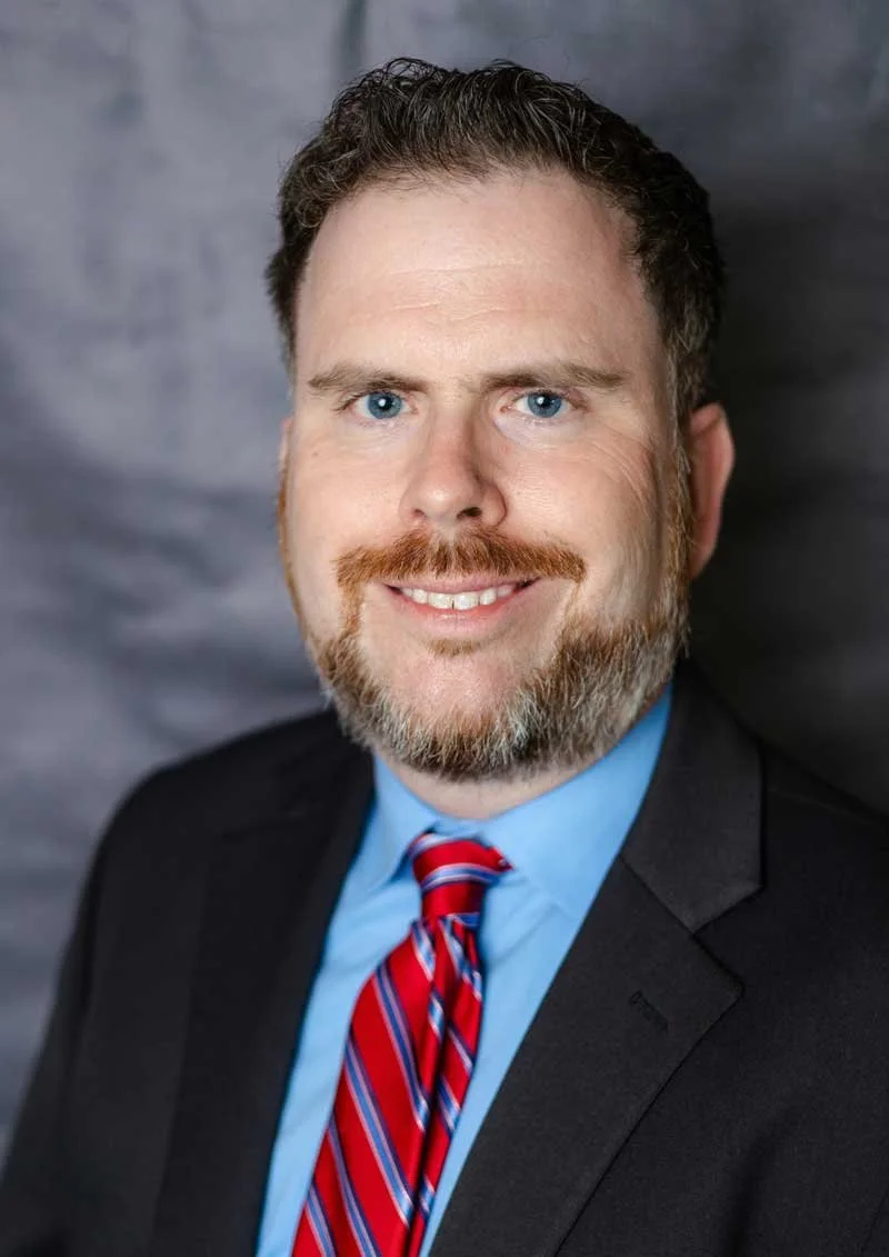A professor at The Cambridge School of Dallas classical Christian school grades 5-12, wearing a black suit, light blue shirt, and a red striped tie, smiling at the camera against a gray background.