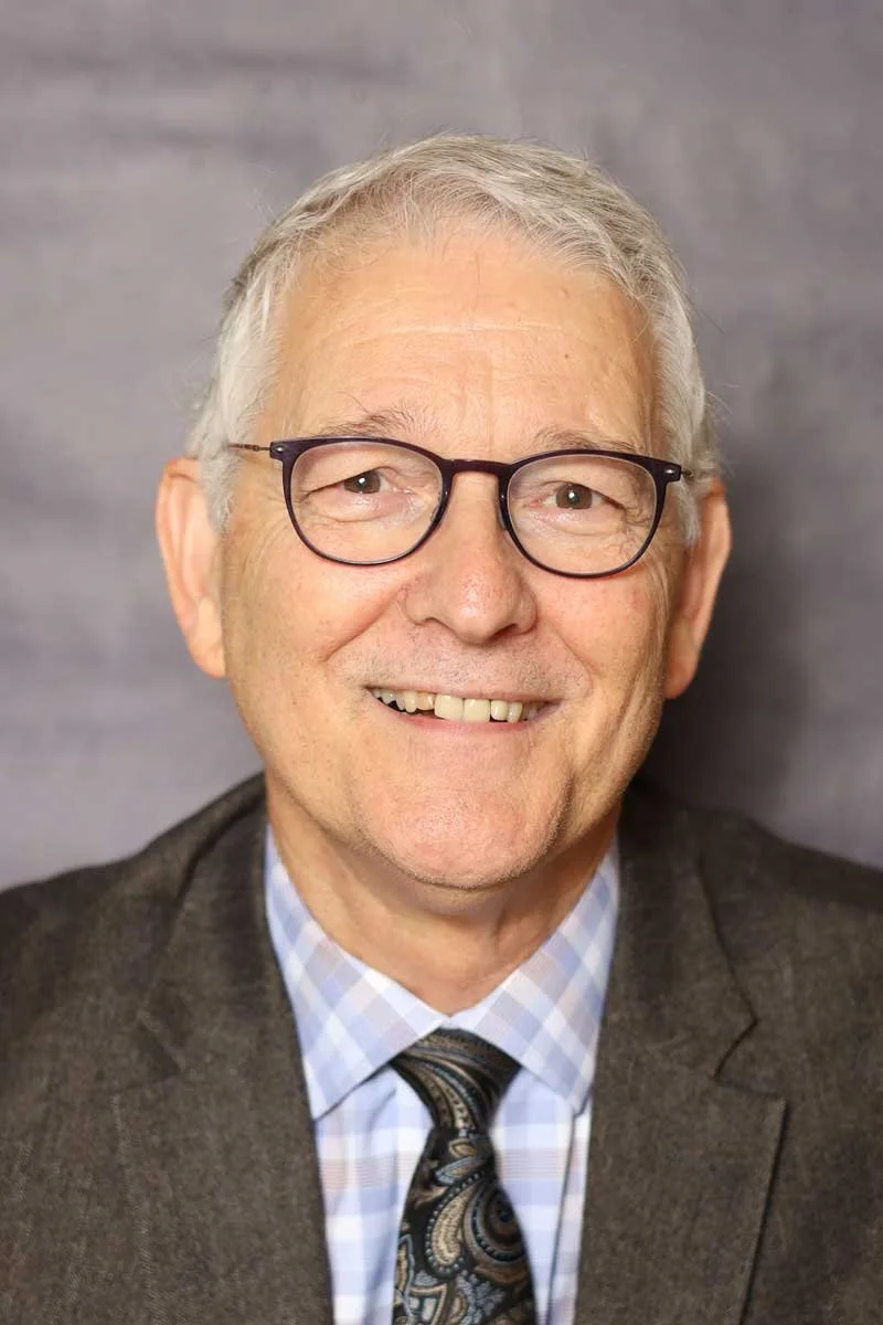 A smiling older man with gray hair, glasses, wearing a suit and tie, against a gray background.