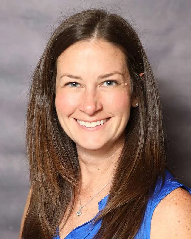 A professor at The Cambridge School of Dallas classical Christian school grades 5-12. A young woman with long brown hair smiling, wearing a blue sleeveless top and a necklace, against a gray background.