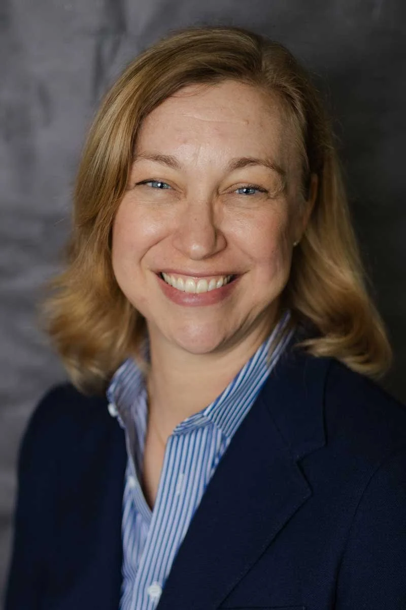 A professor at The Cambridge School of Dallas classical Christian school grades 5-12. A smiling woman with blonde hair, wearing a navy blazer and a blue striped shirt, in front of a gray background.