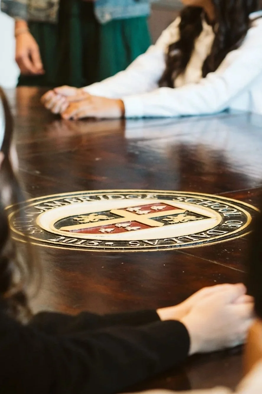 People sitting around a wooden table with a large emblem of the American School of Dubai on it, some hands resting on the table, in a classroom or meeting room setting.