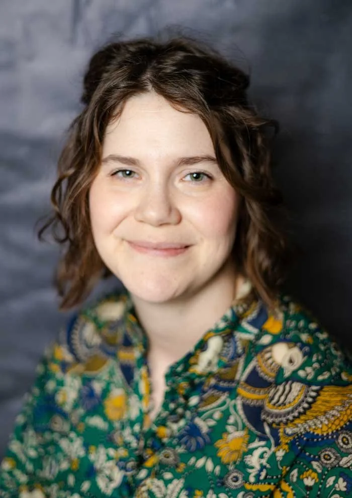 A woman with short, curly brown hair and fair skin, smiling slightly, wearing a colorful, patterned shirt with green, yellow, and blue designs, standing against a dark, blurred background.