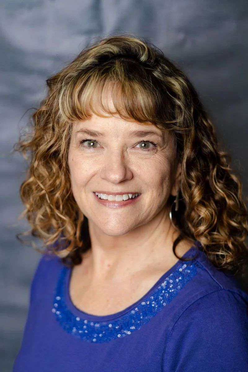 A professor at The Cambridge School of Dallas classical Christian school grades 5-12. Headshot of a woman with curly light brown hair, blue eyes, and a bright smile, wearing a blue top with embellishments.