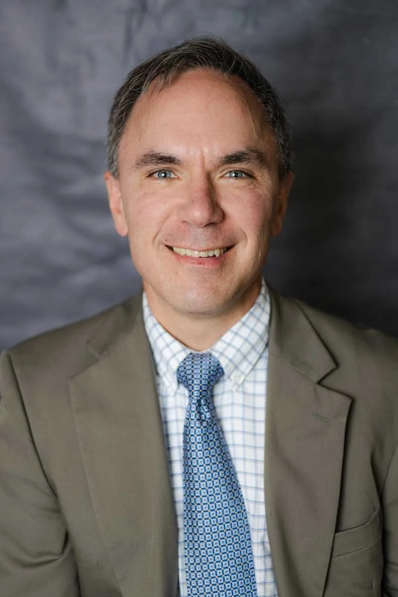 A professor at The Cambridge School of Dallas classical Christian school grades 5-12, smiling, with short brown hair, blue eyes, wearing a tan suit jacket, a checkered dress shirt, and a blue patterned tie, against a gray textured background.