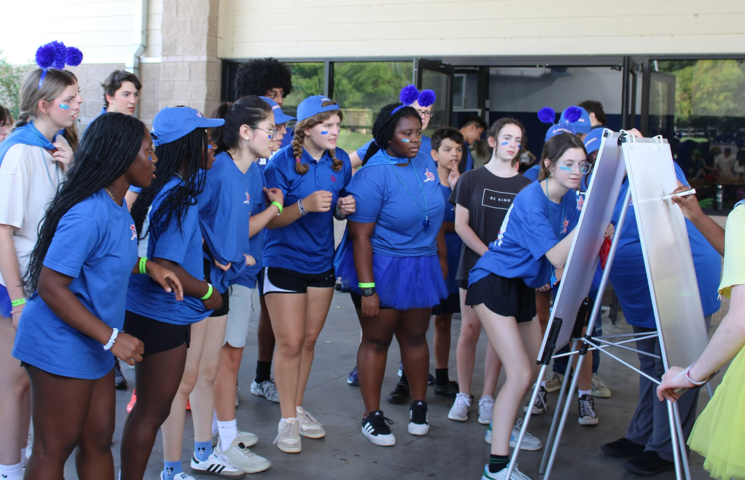 A group of young girls, mostly wearing blue shirts and accessories with purple pom-pom headbands, gather around an outdoor whiteboard with a woman writing on it. Some girls have face paint and colored wristbands, and they are standing outside a building with large windows.