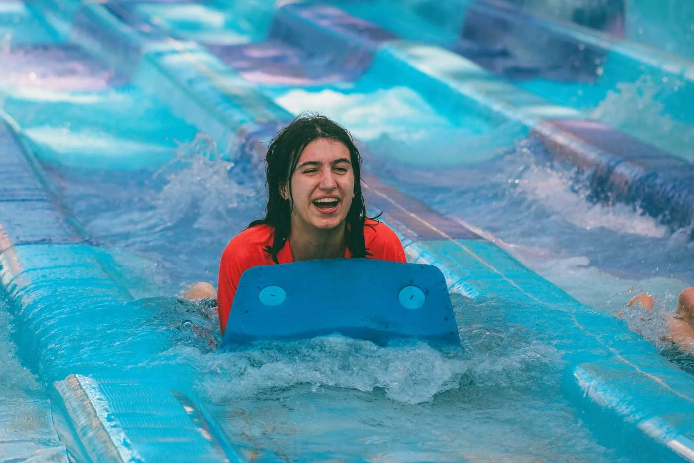 A young woman at a Cambridge School of Dallas retreat in a red shirt is smiling and holding onto a blue water float as she swims in a swimming pool with blue lanes.
