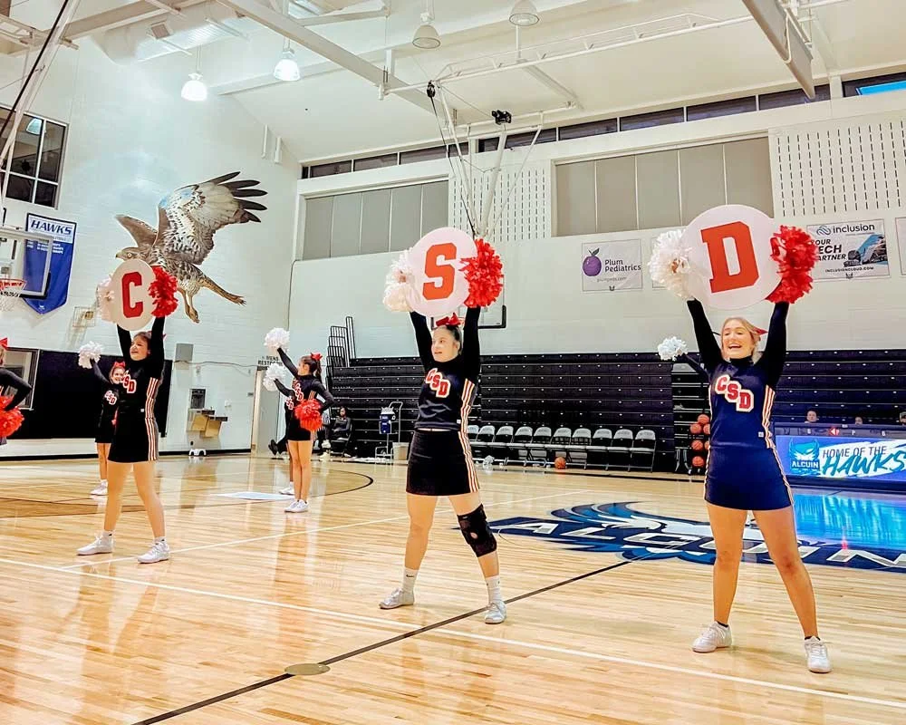 Cheerleaders performing a routine with signs spelling 'C S D' in a gymnasium.