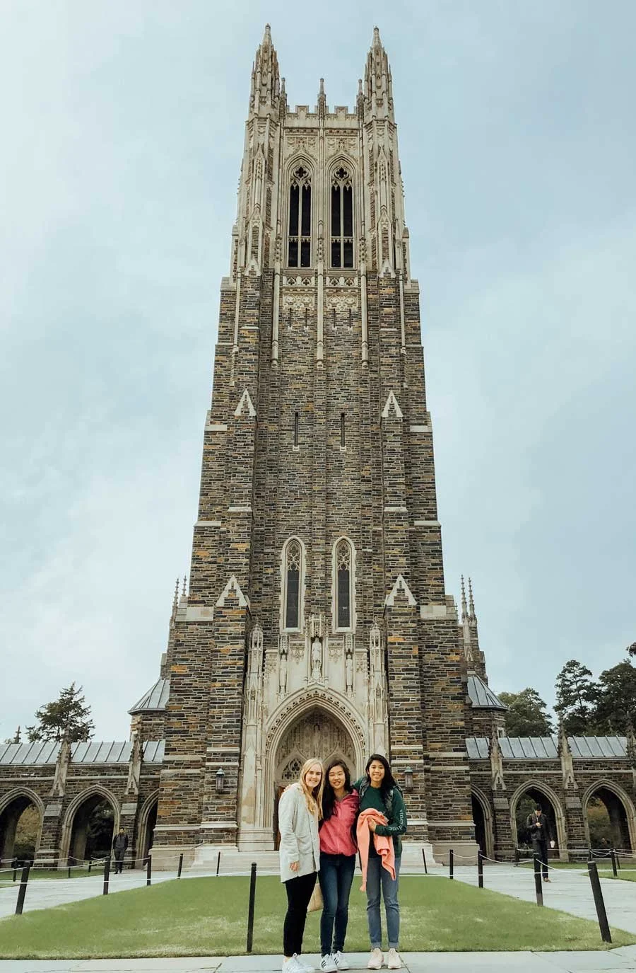 Three women standing in front of a tall, Gothic-style stone tower with arched windows and detailed stonework, on a cloudy day.