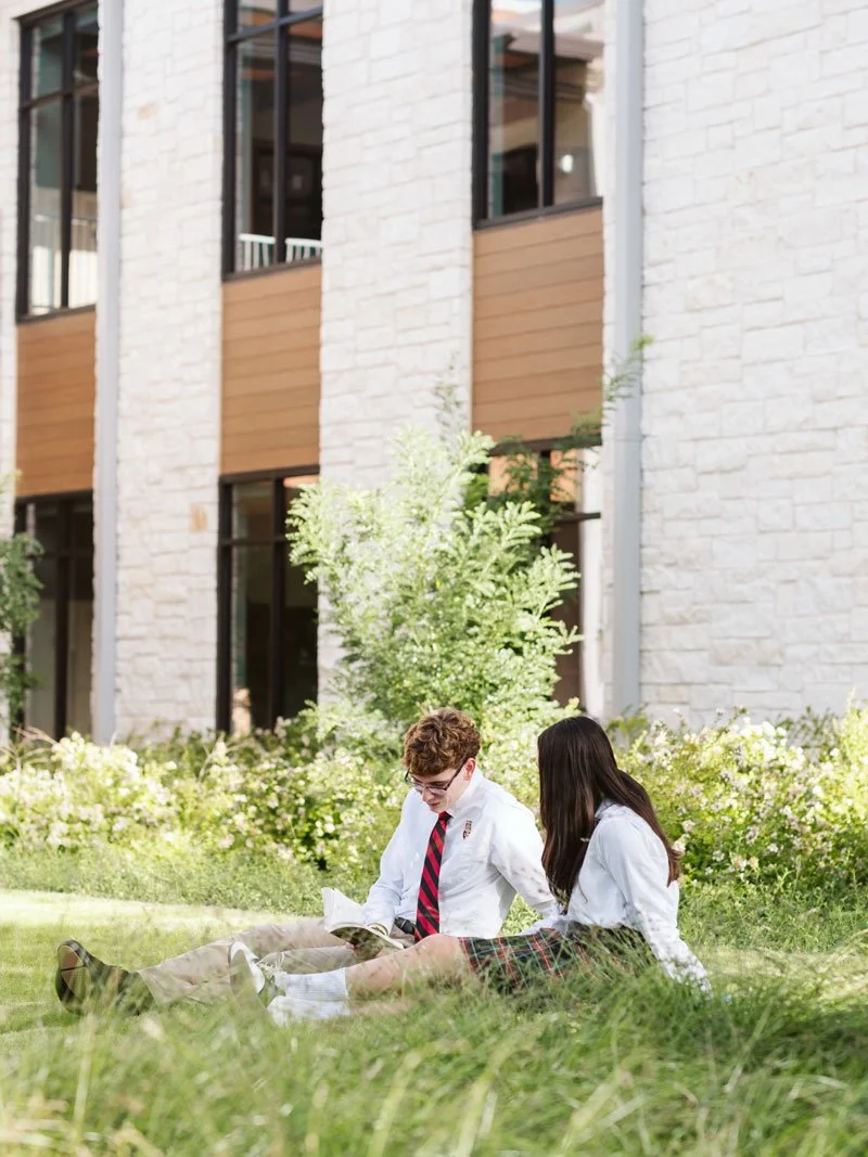 Two students in school uniforms sitting on grass and reading outdoors near a modern building with large windows and light-colored brick walls.
