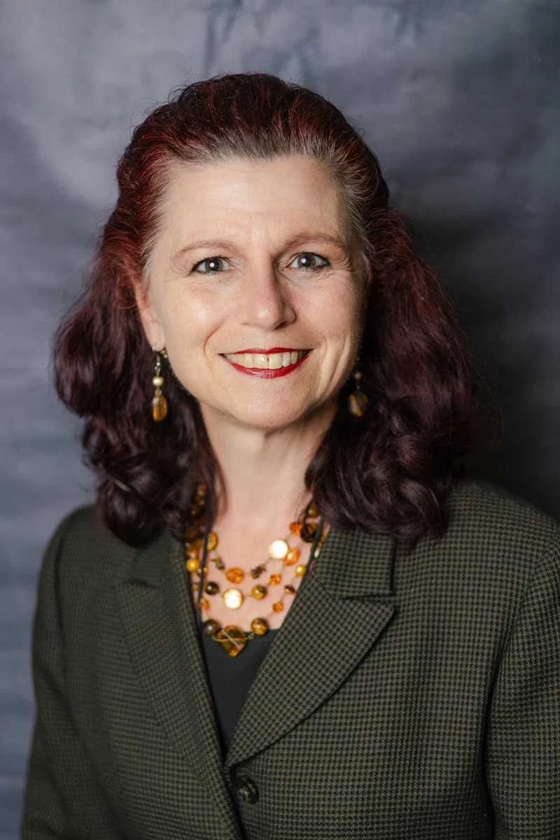 A professor at The Cambridge School of Dallas classical Christian school grades 5-12. A woman with curly, dark red hair and fair skin, smiling in front of a dark, textured background, wearing a dark blazer and jewelry.