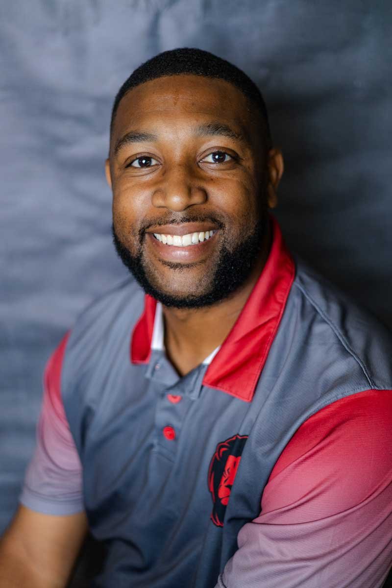 A professor at The Cambridge School of Dallas classical Christian school grades 5-12.. A smiling man with short black hair and beard, wearing a gray and red sports polo shirt with a lion logo on the chest, against a gray background.