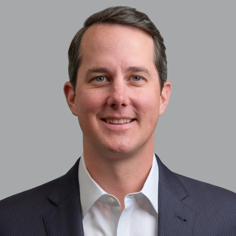 A professional man with short brown hair, wearing a white collared shirt and dark suit jacket, smiling against a gray background.