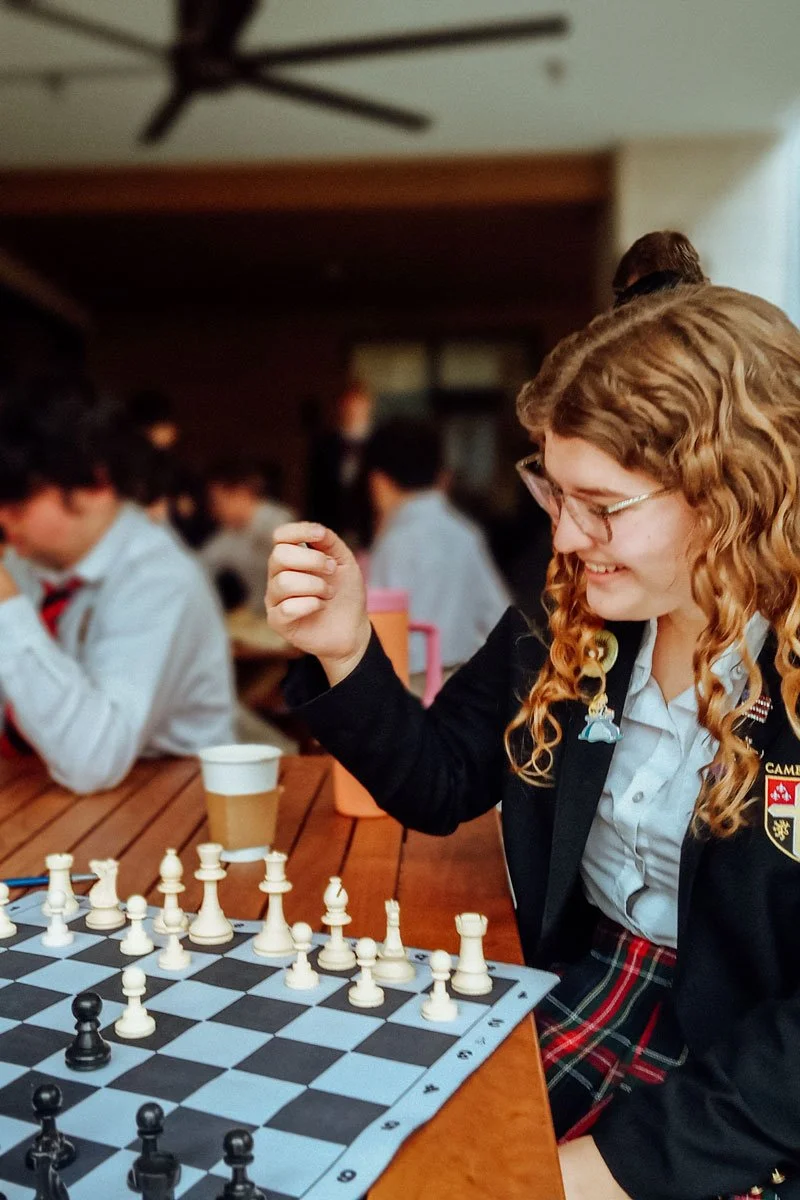 Female student at the Cambridge School of Dallas with curly hair and glasses playing chess in a classroom or club setting, with other students in the background.