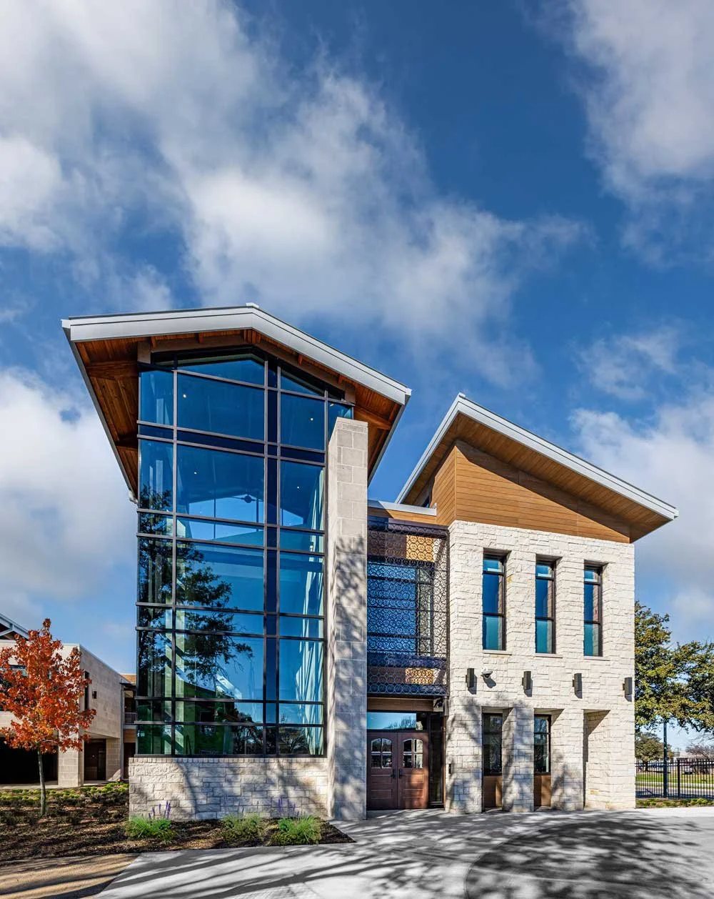 Modern multi-story house with large glass front on the left and stone exterior walls, surrounded by trees and a blue sky with clouds.