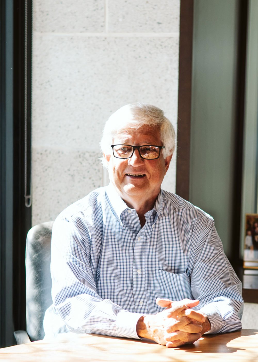 An elderly man with gray hair and glasses sitting at a table, smiling and talking in a bright office with sunlight coming through the window.