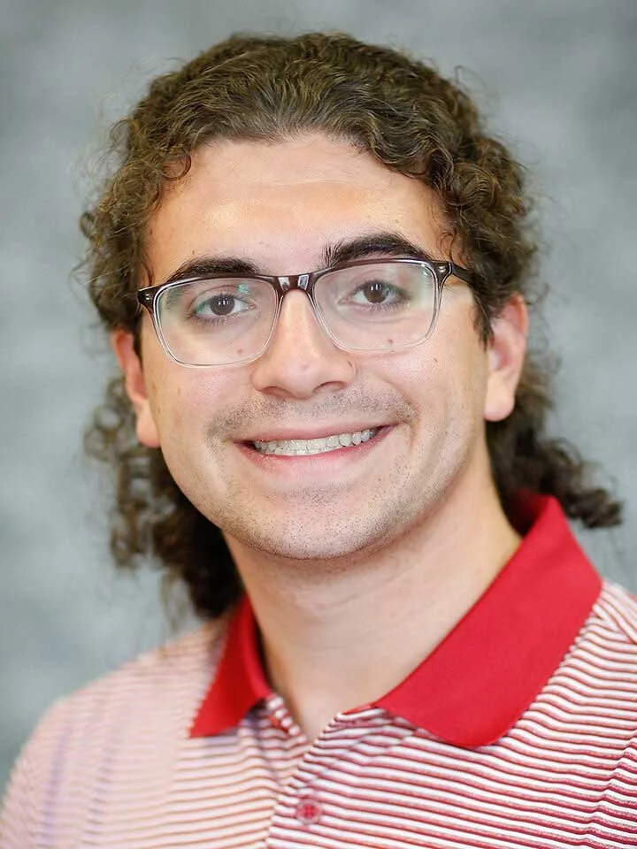 Close-up portrait of a young man with curly brown hair, glasses, and a friendly smile, wearing a red and white striped polo shirt with a red collar.