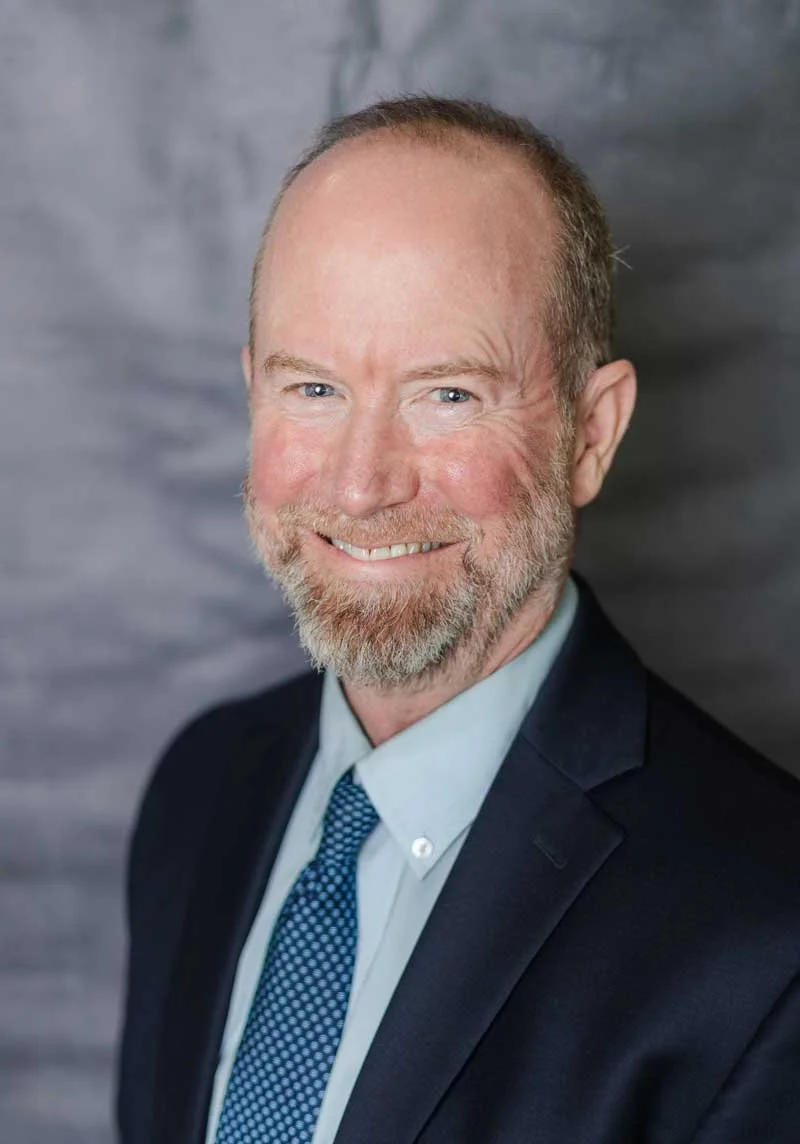A professor at The Cambridge School of Dallas classical Christian school grades 5-12, wearing a dark suit, light blue shirt, and blue patterned tie, against a gray background.
