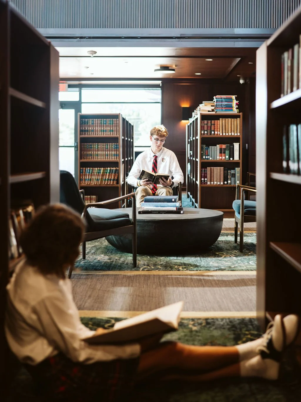 A young person in school uniform reading a book in a library, seated on a table, with another person lying on the floor reading a book in the foreground.