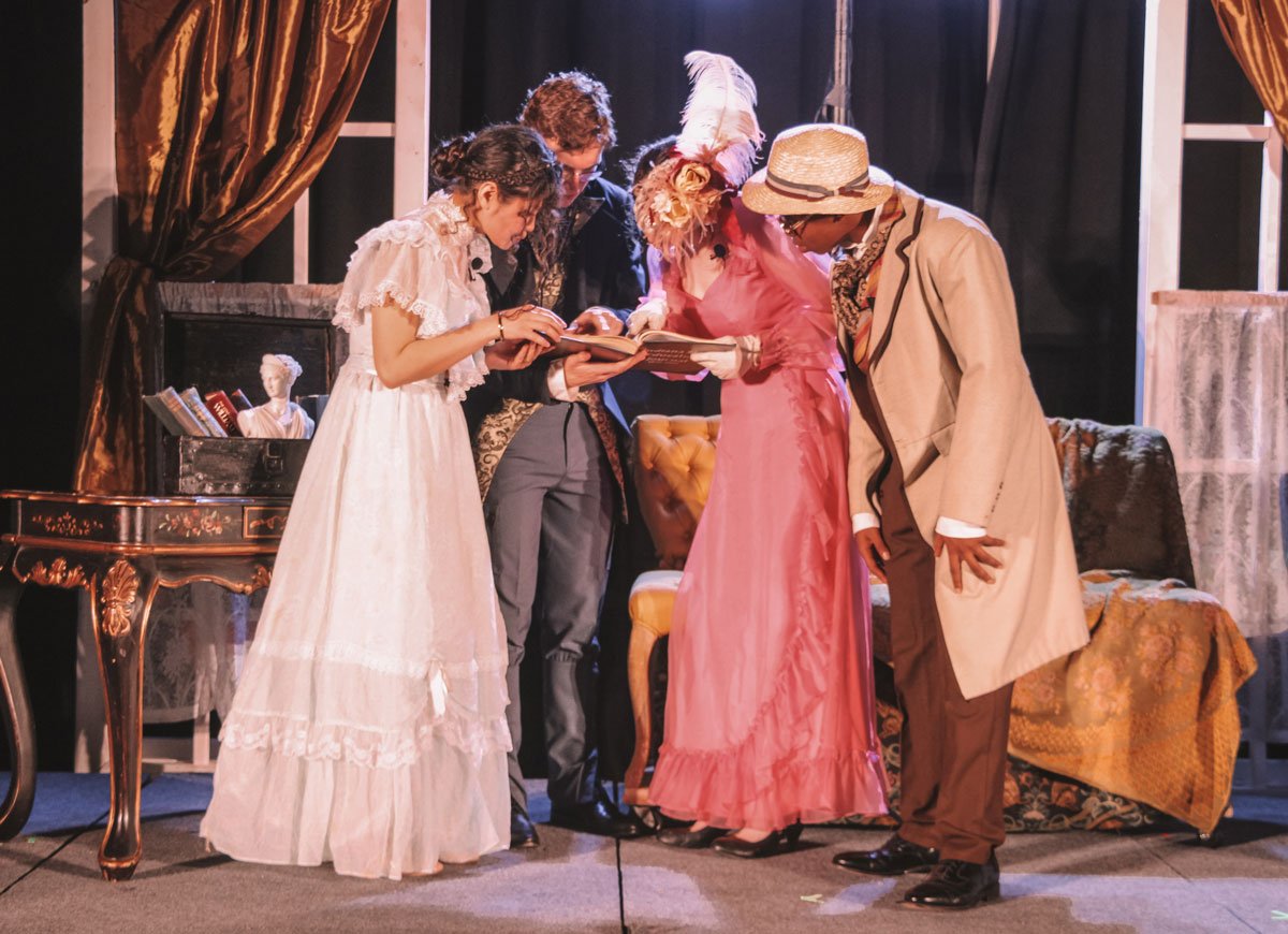 Five people in period costumes including dresses, a coat, and a hat, gathered around reading a book on stage with vintage furniture and drapery in the background, performing The Importance of Being Earnest at the Cambridge School of Dallas.