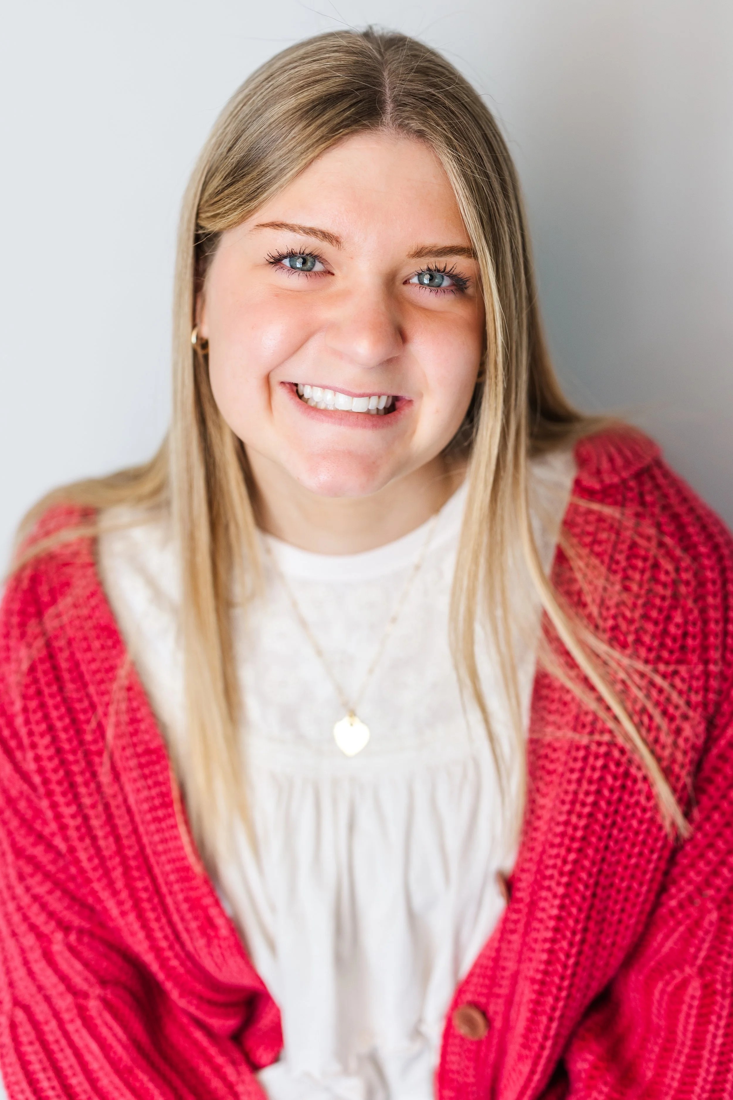 Young woman with blonde hair and blue eyes smiling, wearing a red knitted cardigan and a white blouse, with a gold heart-shaped necklace.