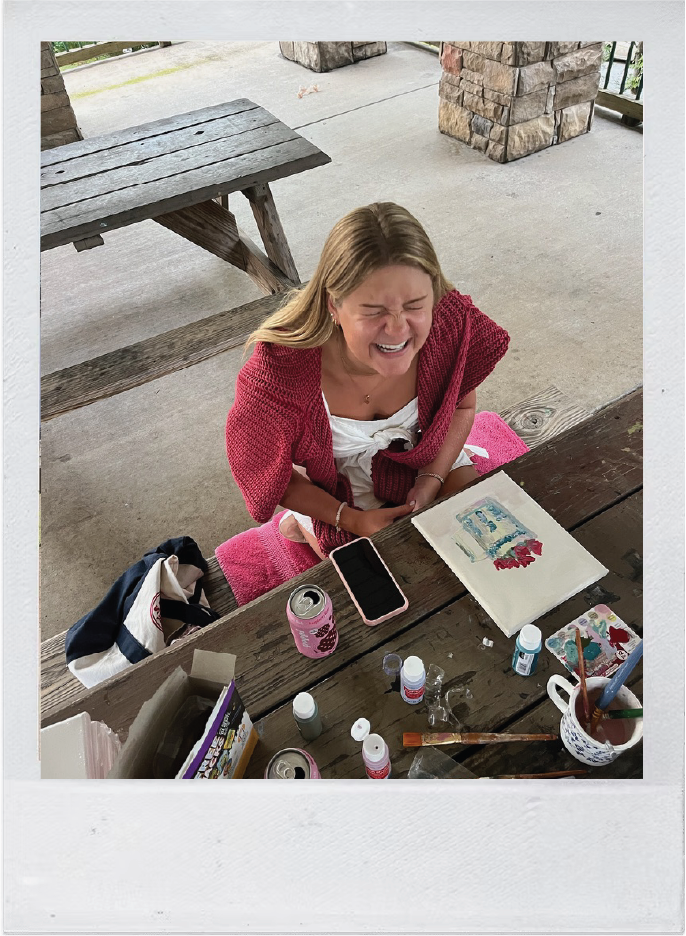 A young woman sitting at a wooden picnic table outdoors, smiling and laughing with her eyes closed, surrounded by art supplies and a watercolor painting.