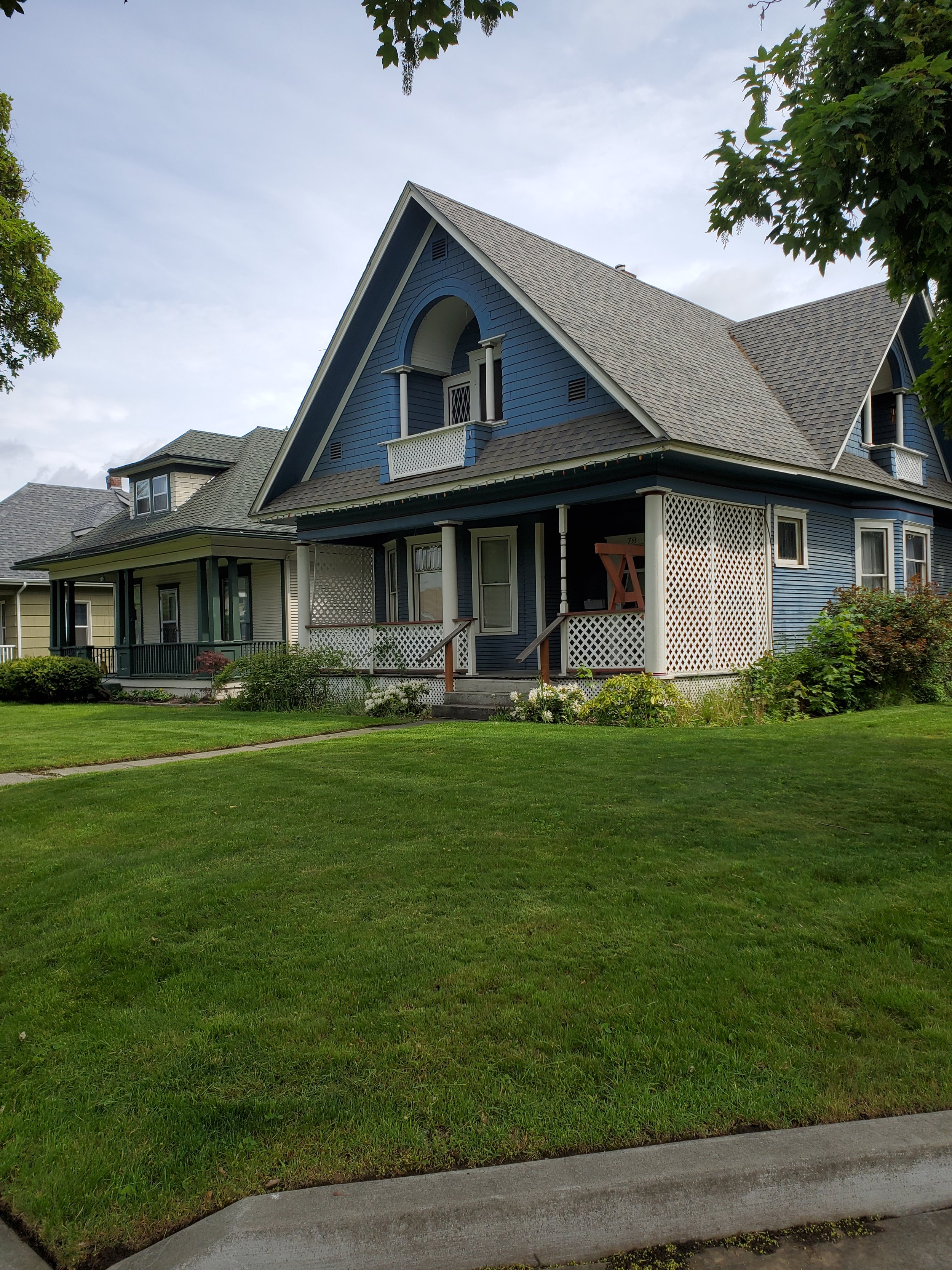 A large blue Victorian-style house with a front porch, decorative latticework, and a steep pitched roof, surrounded by green lawn and trees.