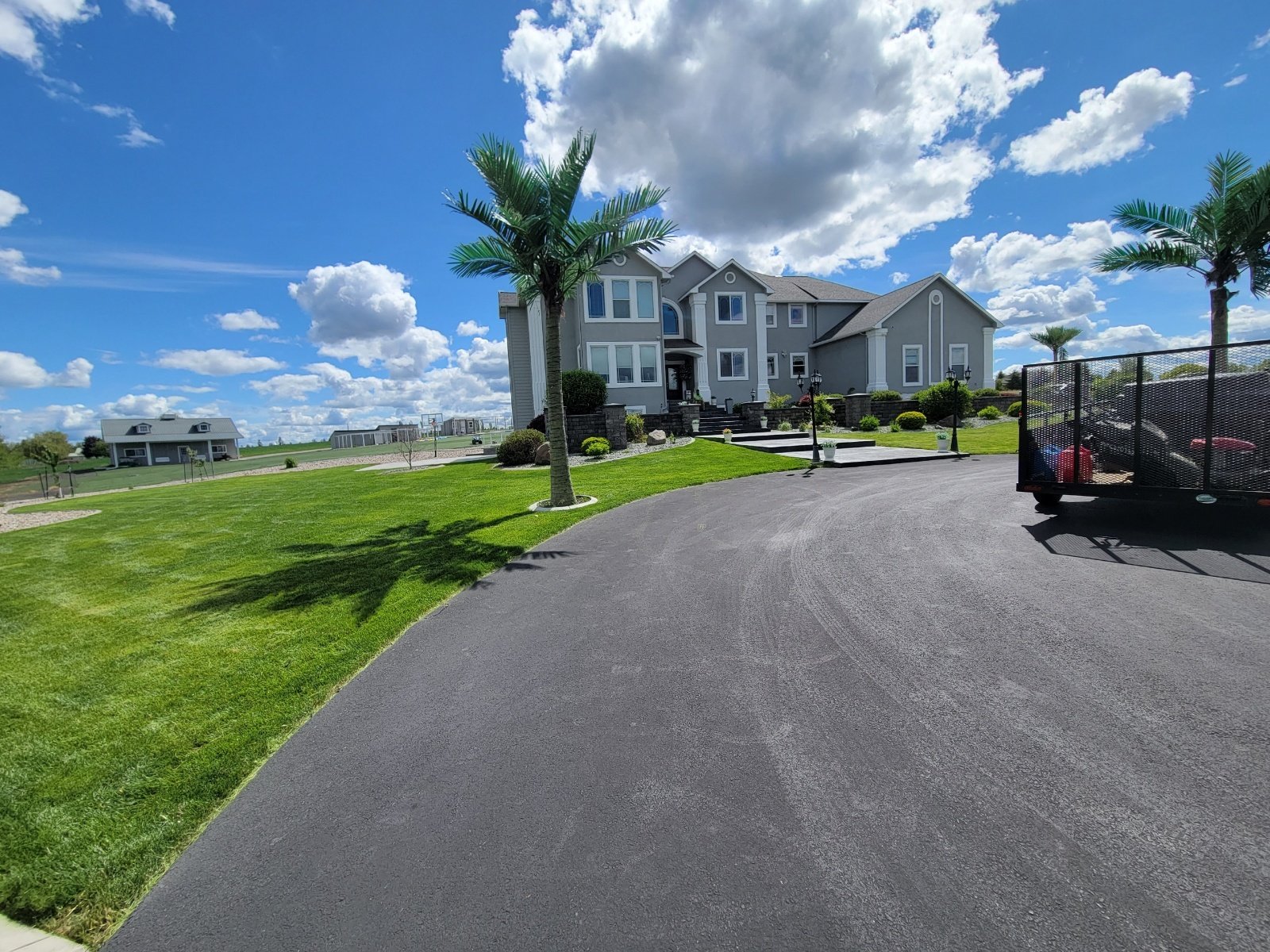 Large gray house with multiple stories, front steps, and a landscaped yard with palm trees, under a partly cloudy blue sky.