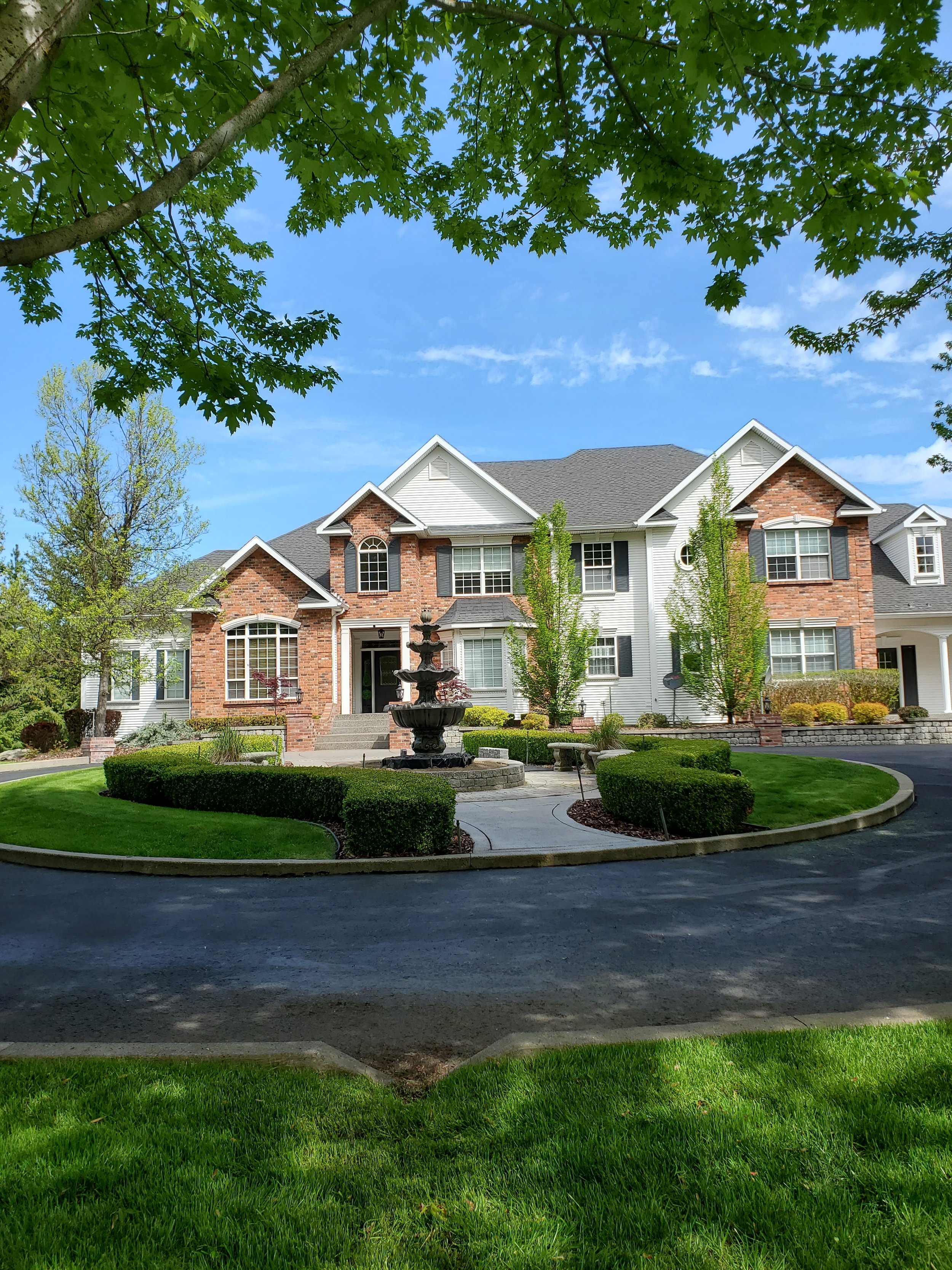 Large two-story house with brick and white siding, front yard with green lawn, trees, and a fountain, under a blue sky with some clouds.