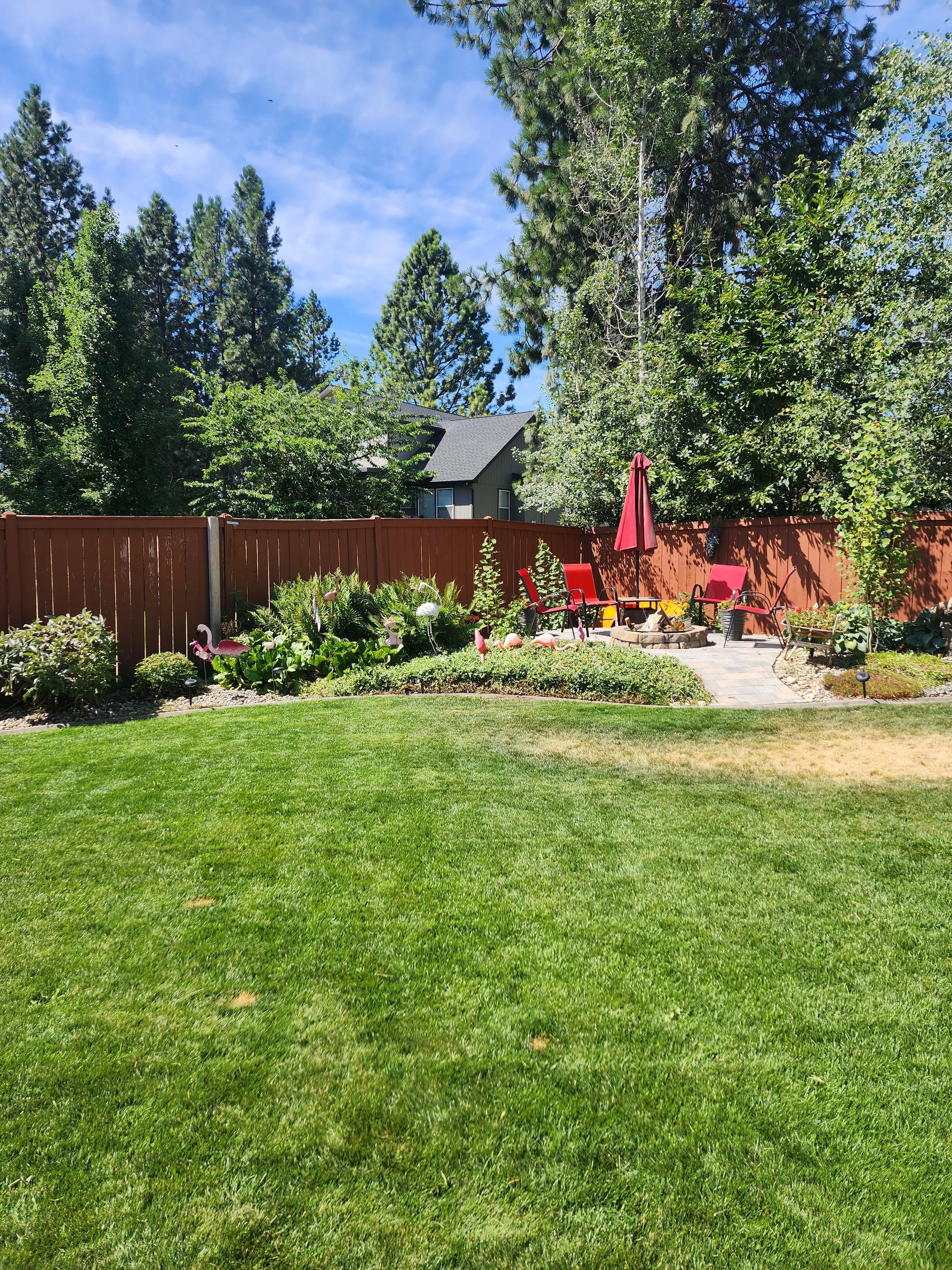 A backyard with a lush green lawn, a flower garden with pink flamingo decorations, a stone patio with red chairs, a red umbrella, and surrounded by tall trees and a wooden fence.