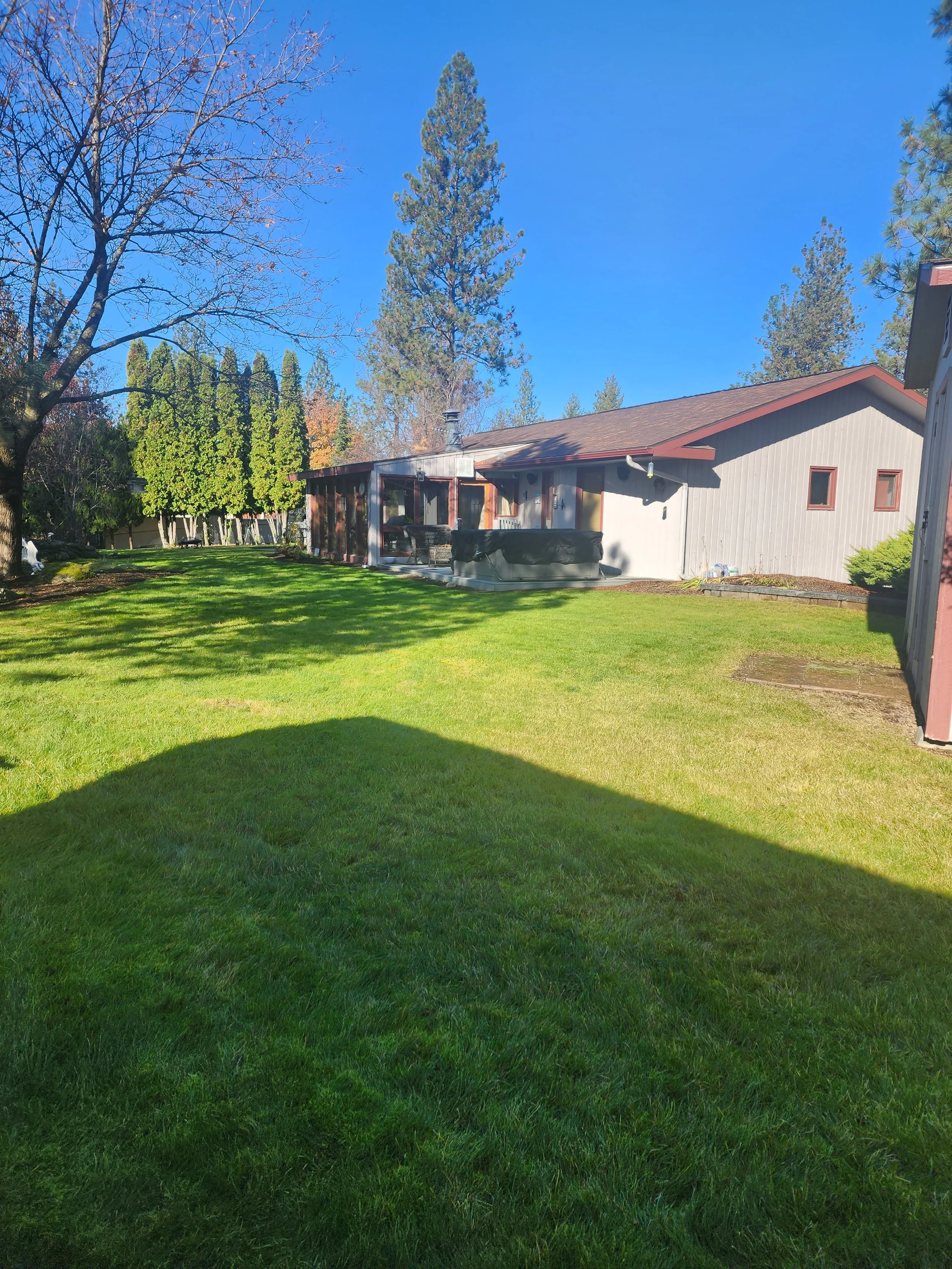 A backyard with green grass, trees, a house with a brown roof, and a patio area with furniture, under a clear blue sky.