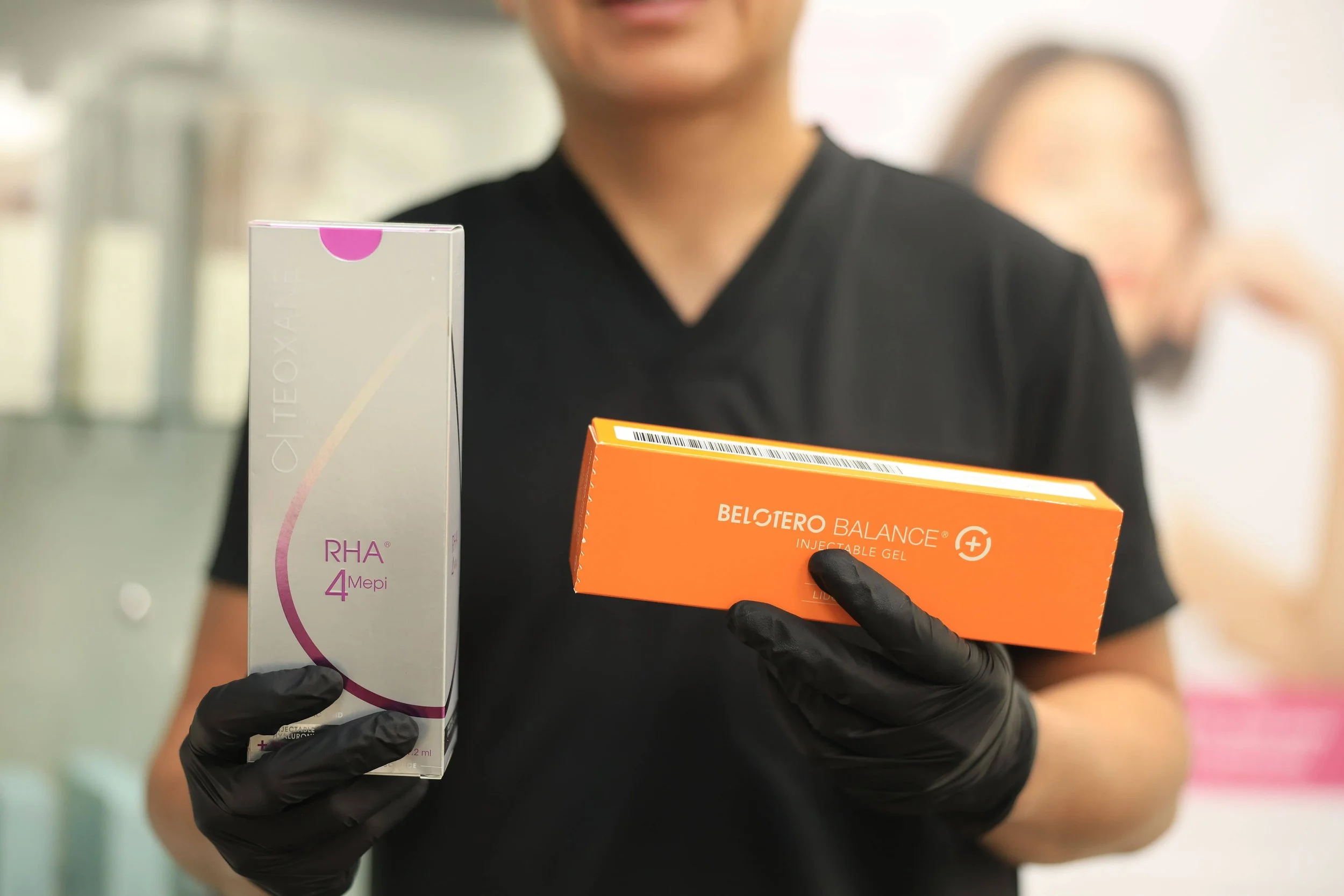 Injector in black gloves holding boxes of Teoxane RHA 4 and Belotero Balance dermal filler for a filler correction consultation.
