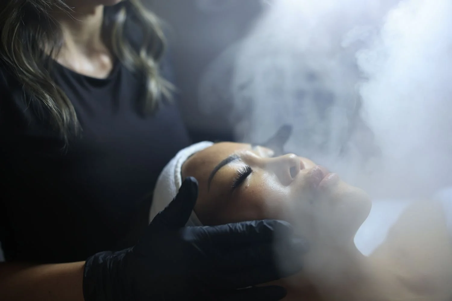 A woman receives a facial treatment from an esthetician, with steam or vapor around her face, in a spa or skincare clinic setting.