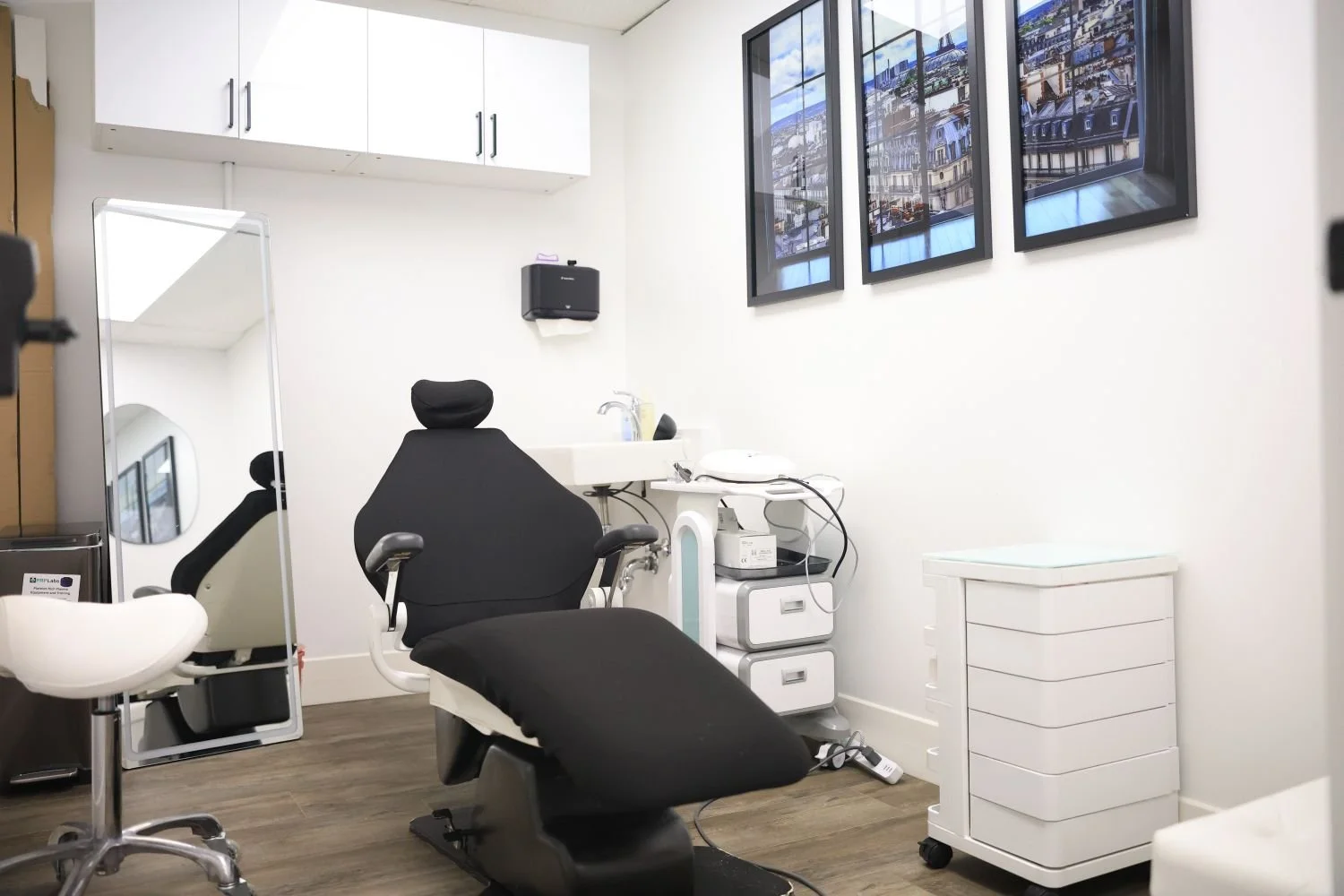 Modern dental examination room with a black dental chair, white walls, framed cityscape photographs, and medical equipment.