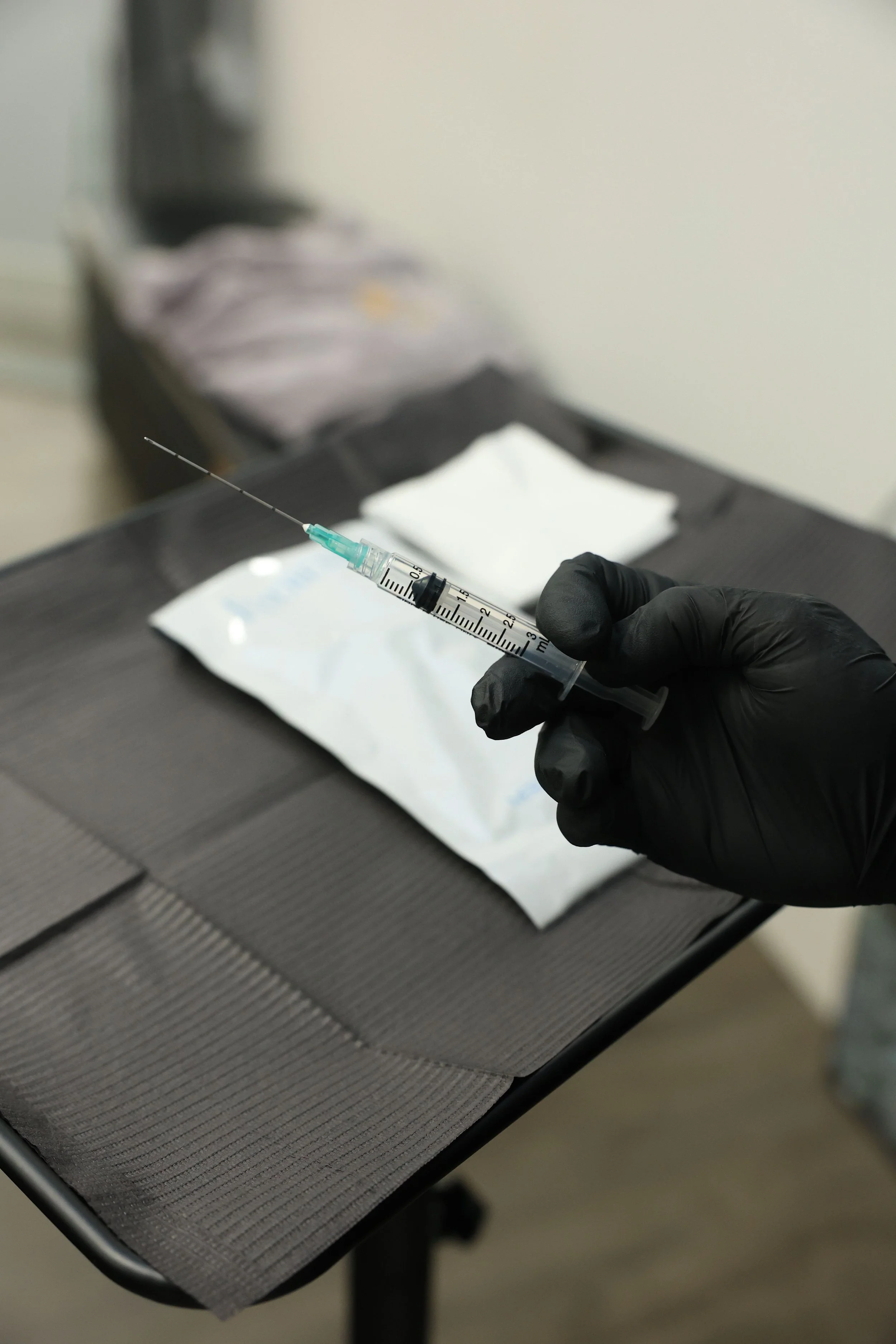 A gloved hand holding a syringe, with a medical sterile package and gauze on a black surface in the background.