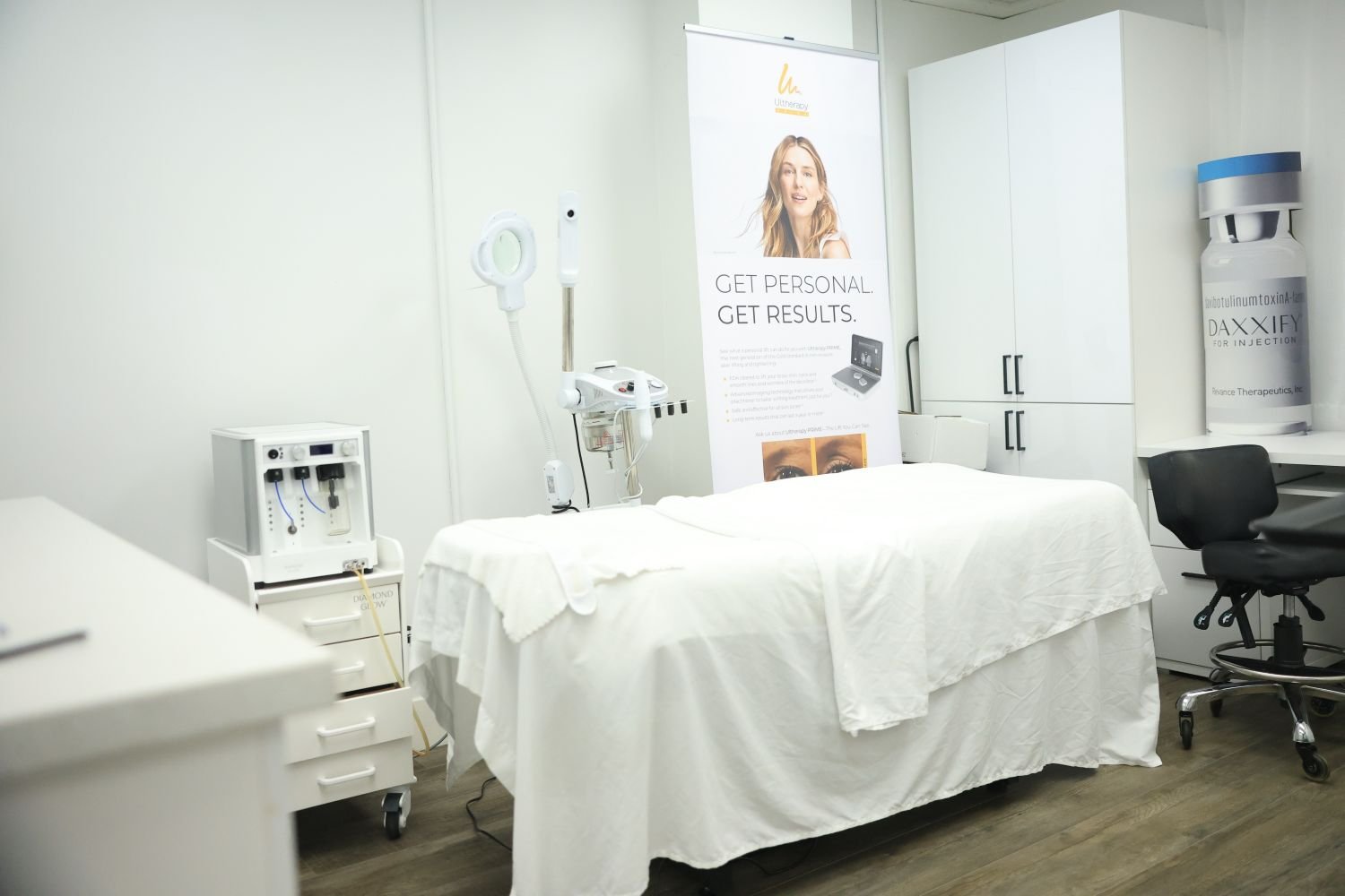 Medical examination room with a bed covered in white sheets, medical equipment, a black chair, white cabinets, and a large banner promoting personalized skincare results.
