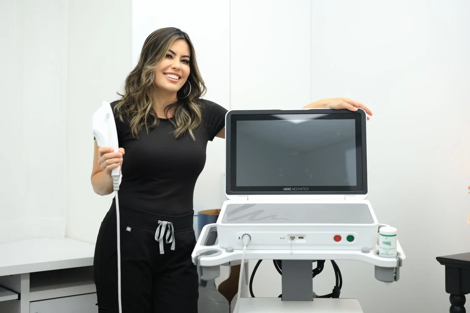 Woman smiling in a medical or aesthetic setting, holding a handheld ultrasound device next to a large white medical machine with a screen and control buttons.