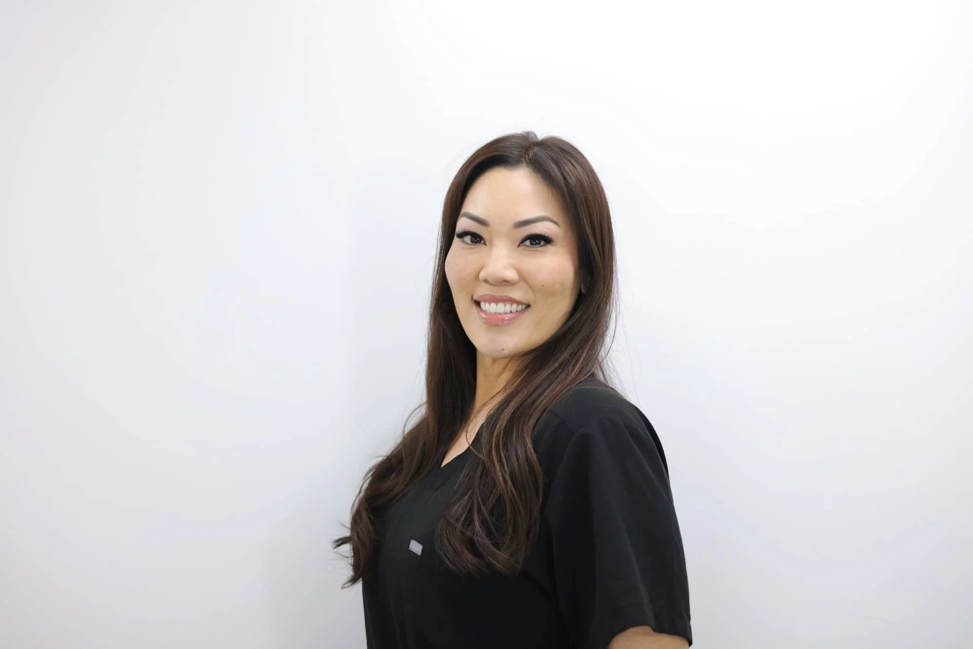A smiling woman with long brown hair, wearing a black shirt, standing against a plain white background.