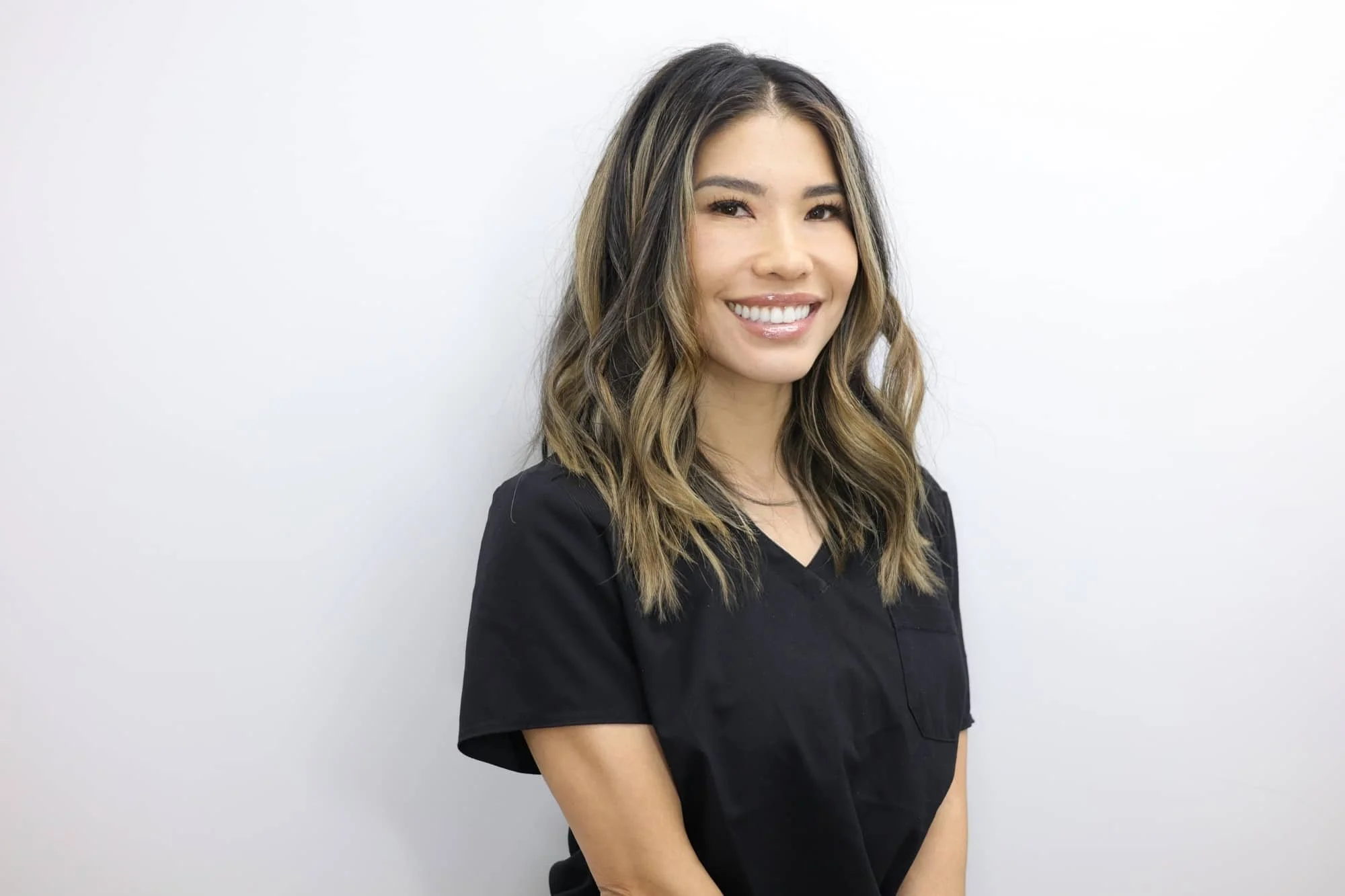 Portrait of a smiling woman with wavy brown hair wearing a black top, standing against a white background.