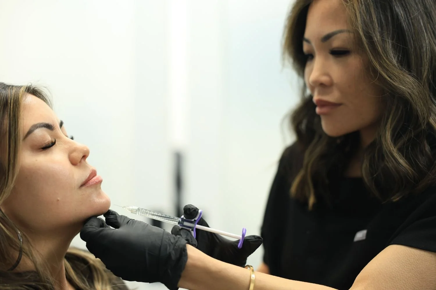 A woman is lying with her eyes closed as a professional in black gloves administers a cosmetic procedure with a syringe on her face, while another woman looks on.