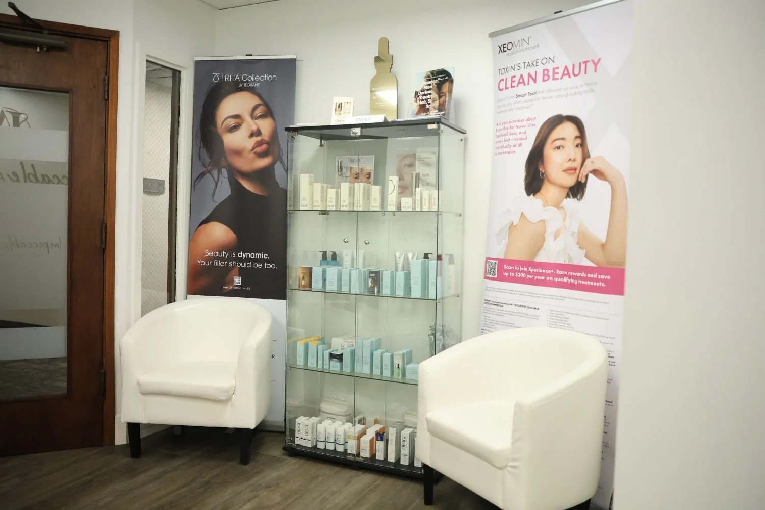 A waiting area inside a beauty clinic with two white armchairs, a glass display cabinet filled with skincare products, and promotional posters on the wall featuring women with flawless skin and promoting skincare treatments.