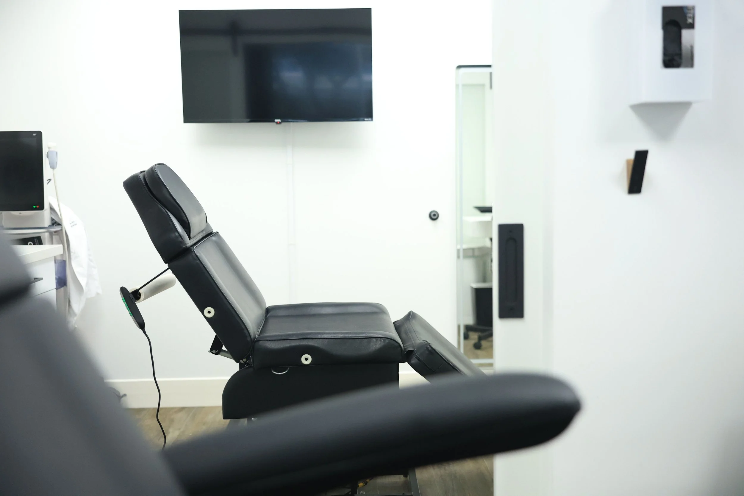 A clinical examination room featuring a black examination chair, a mounted television, and some medical equipment.