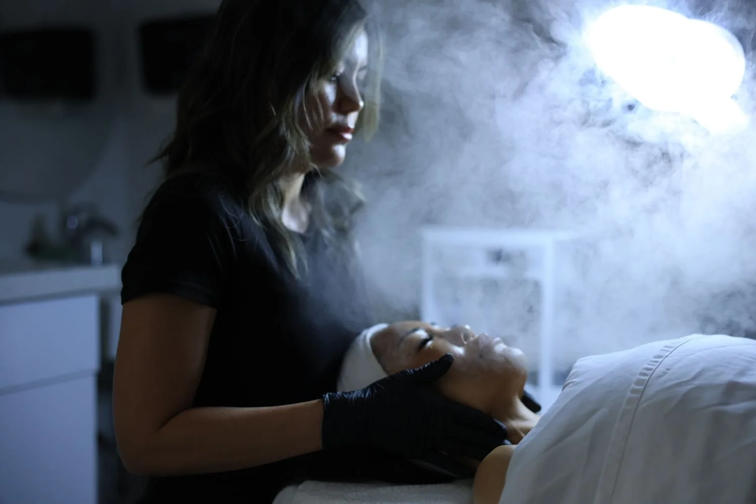 A woman in black gloves tending to a person lying on a bed in a dimly lit room with steam or smoke in the air.