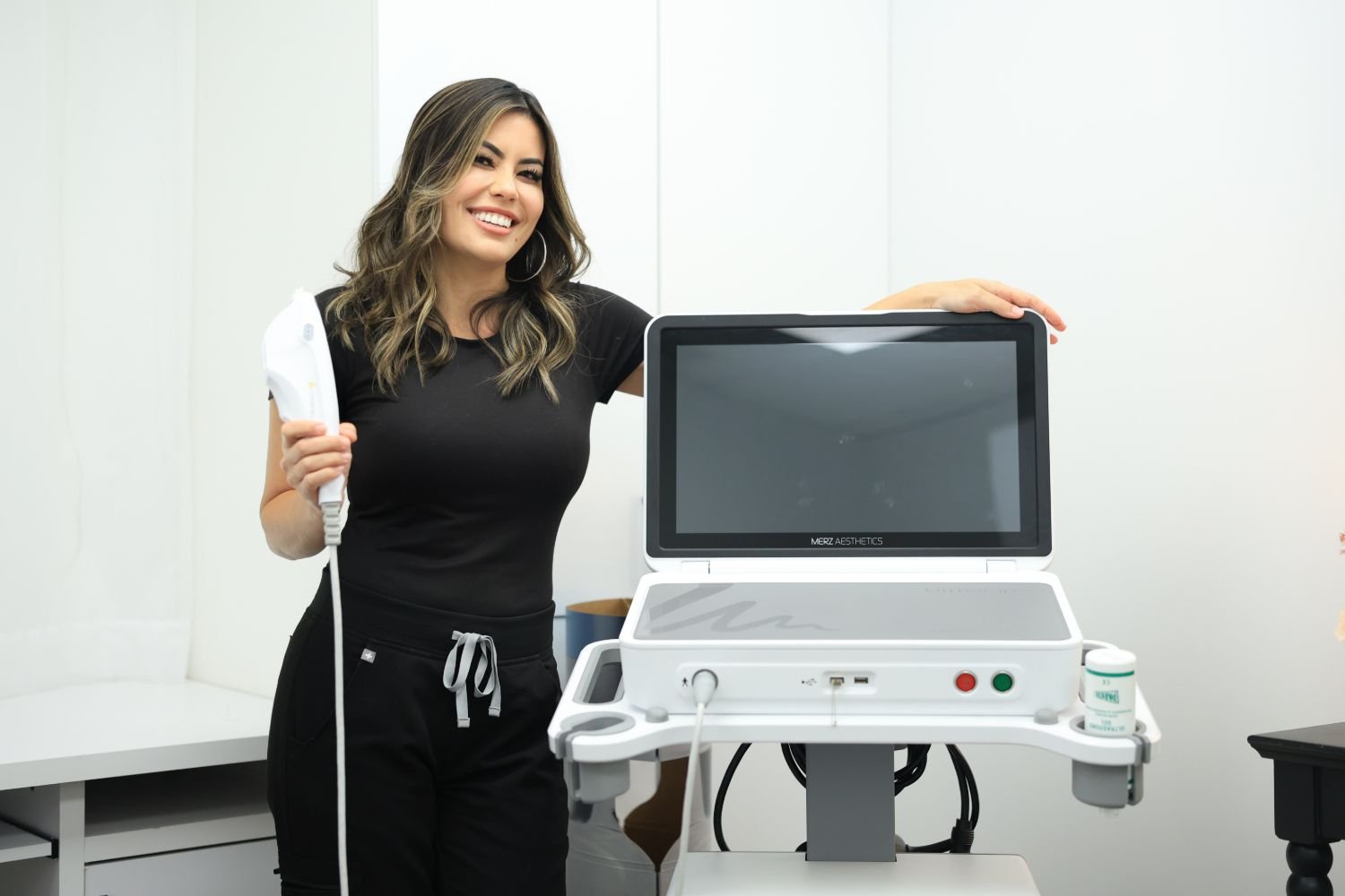 A woman in black scrubs smiling and holding a medical device next to a large medical machine with a screen in a clinical setting.