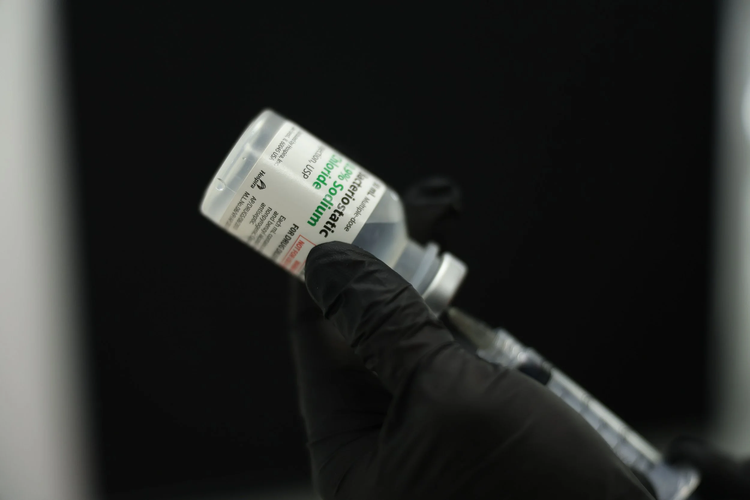 A person wearing black gloves holding a vial of emergency use authorized COVID-19 vaccine against a black background.